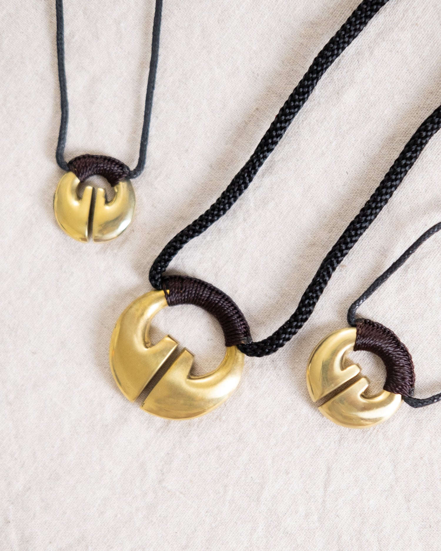 An Ifugao Lingling-o necklaces featuring a traditional brass Lingling-o pendant in varying sizes, shaped like a stylized open hoop with a central cut. The pendants are attached to dark braided or corded strings. Ifugao Lingling-O Necklace Jewelry | Narra Studio