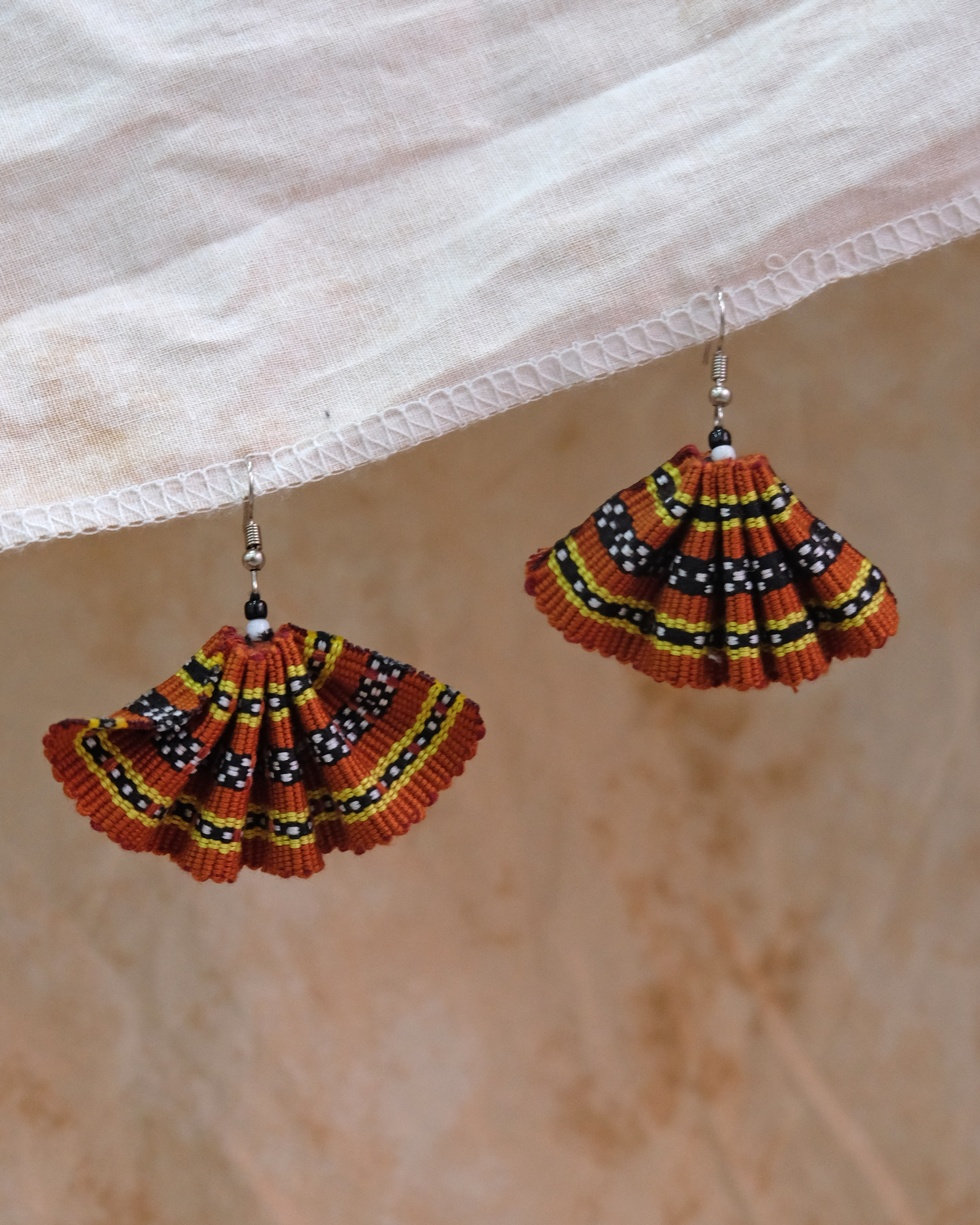 A fan handwoven earring in ochre tones, featuring intricate Kalinga-inspired beadwork and thread patterns in black, yellow, and white on a burnt orange base.