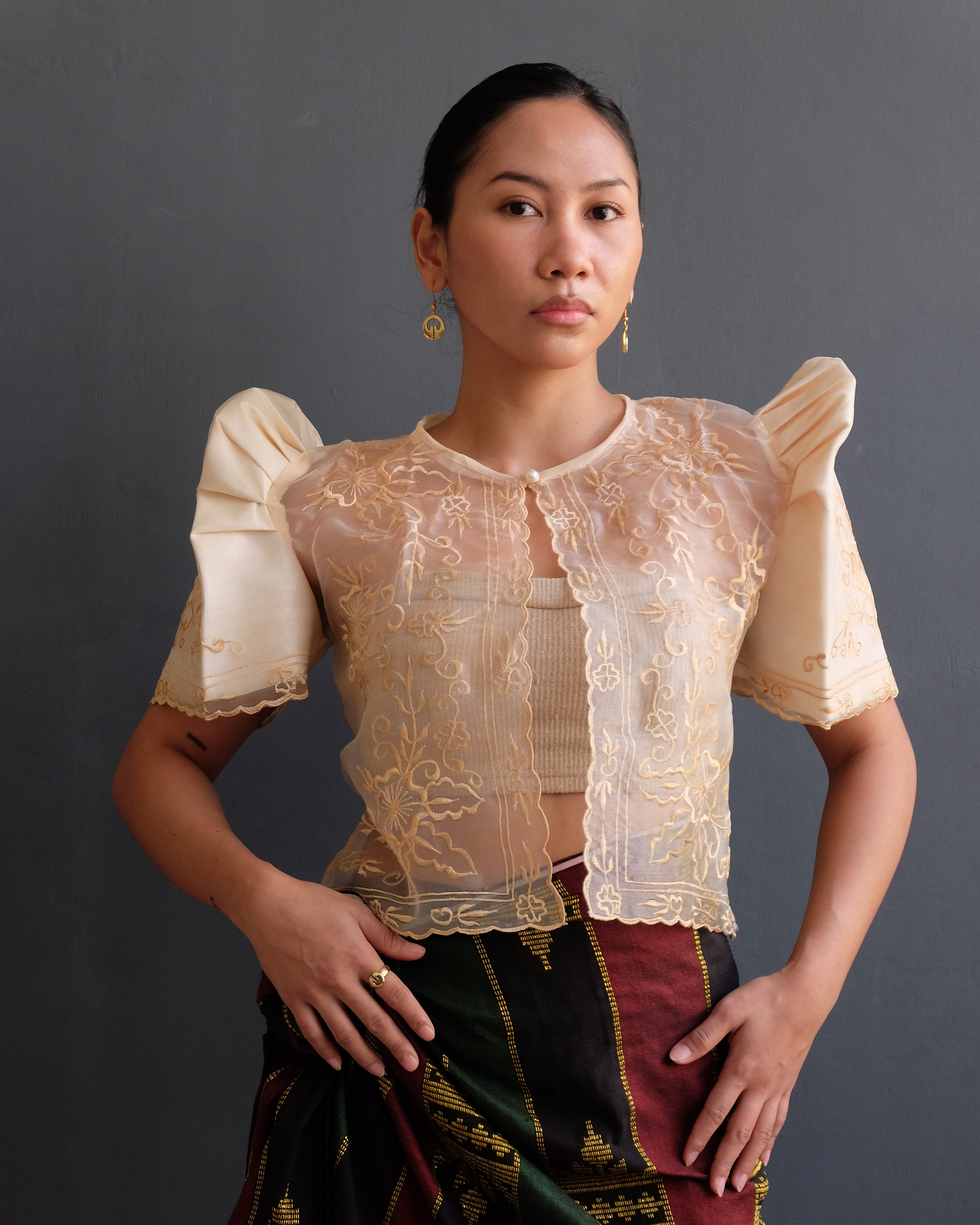 A woman wearing a cream Filipiniana bolero blouse with butterfly sleeves and delicate embroidery on sheer organza fabric, paired with a traditional woven skirt.