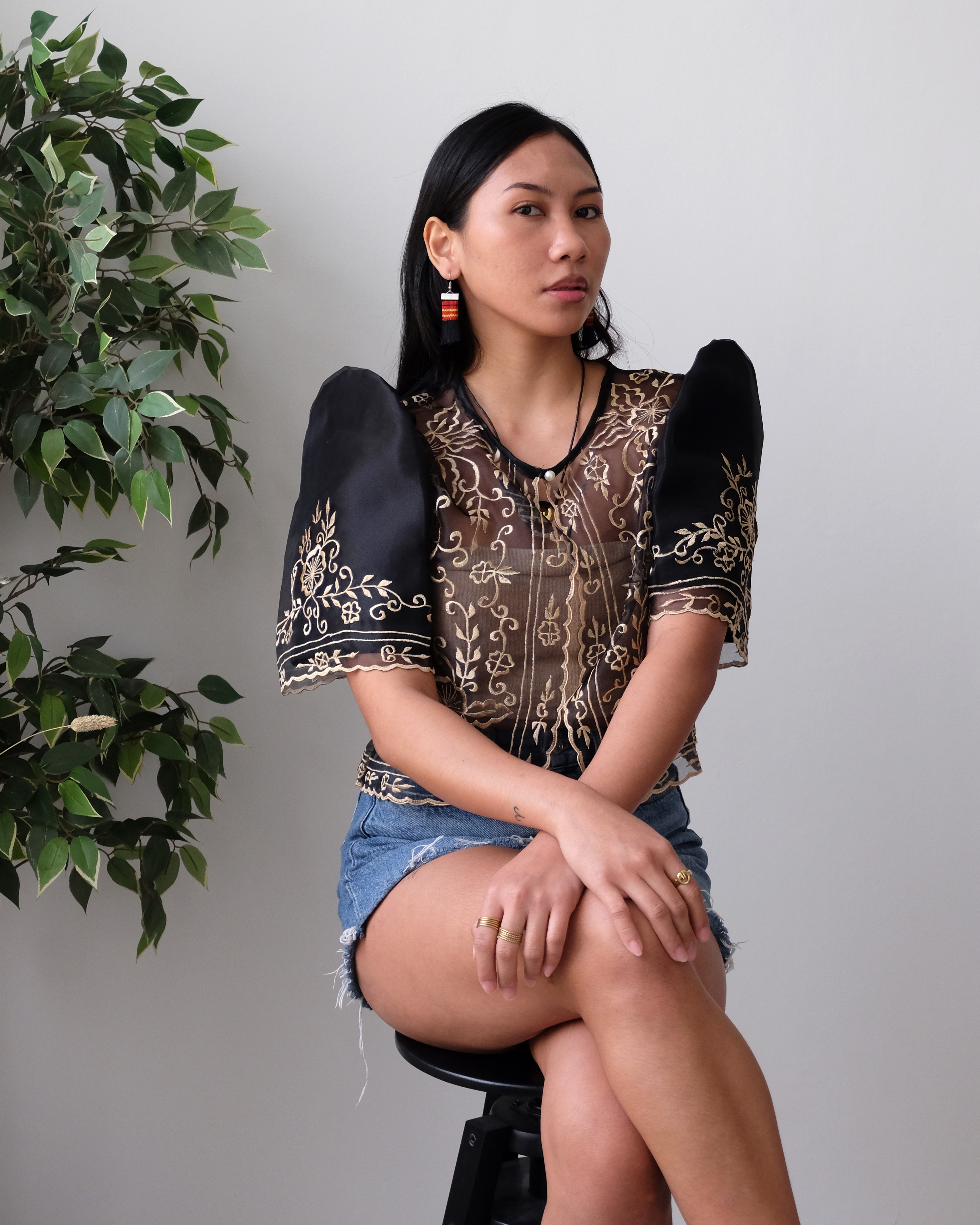 A woman wearing a Black Filipiniana bolero with traditional butterfly sleeves, featuring embroidery on organza fabric, paired with denim shorts and statement earrings.