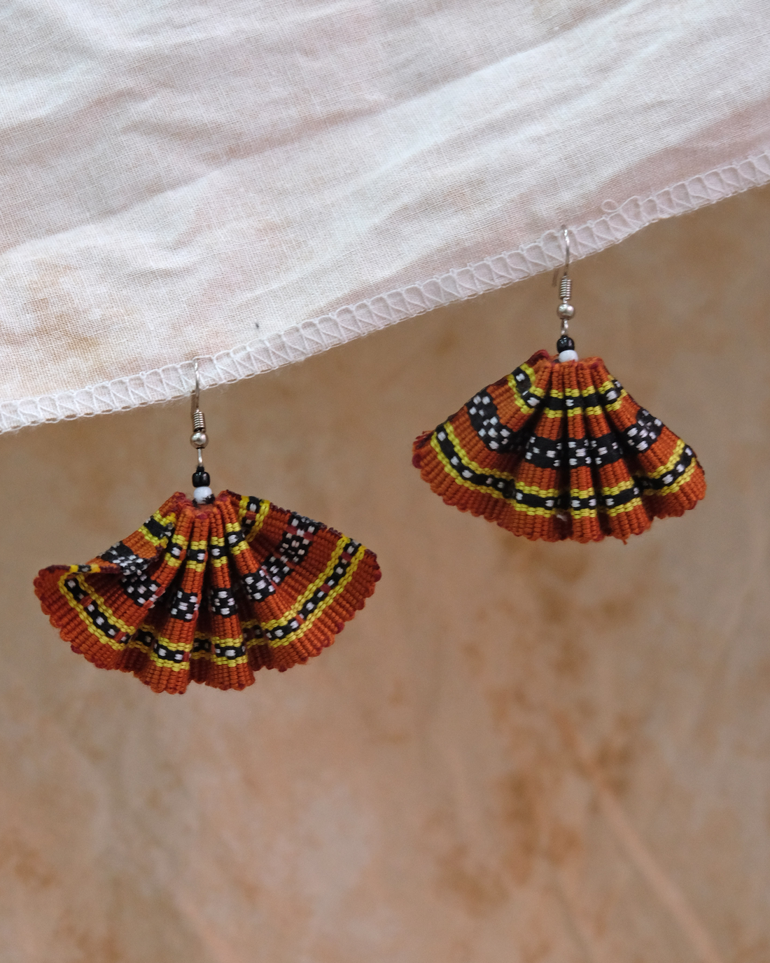 A fan handwoven earring in ochre tones, featuring intricate Kalinga-inspired beadwork and thread patterns in black, yellow, and white on a burnt orange base.