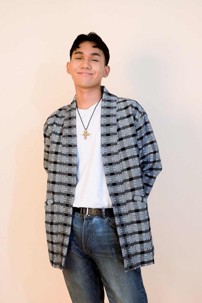 Person wearing a handwoven black Binakol jacket with a grid pattern over a white shirt and blue jeans in a neutral background.