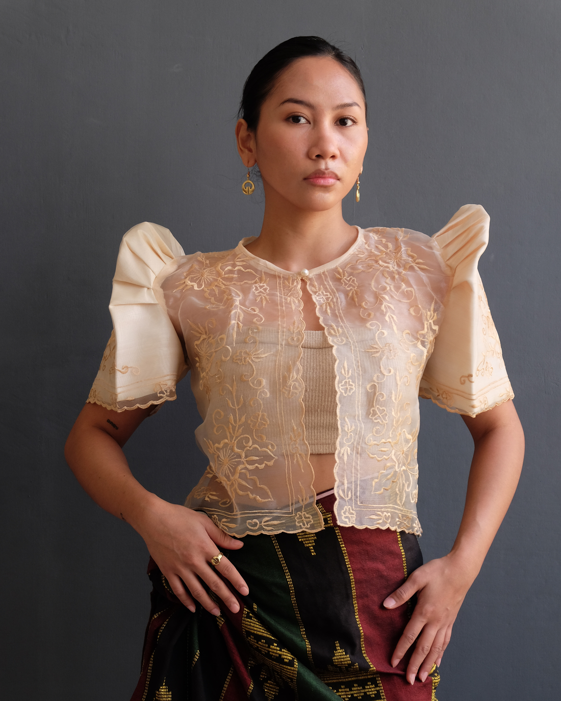 A woman wearing a cream Filipiniana bolero blouse with butterfly sleeves and delicate embroidery on sheer organza fabric, paired with a traditional woven skirt.