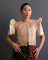 A woman wearing a cream Filipiniana bolero blouse with butterfly sleeves and delicate embroidery on sheer organza fabric, paired with a traditional woven skirt.