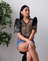 A woman wearing a Black Filipiniana bolero with traditional butterfly sleeves, featuring embroidery on organza fabric, paired with denim shorts and statement earrings.
